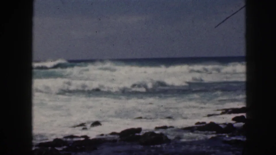 1959 Hawaii home-movie frame of rough surf breaking beyond dark rocks, seen through heavy vertical edge framing.