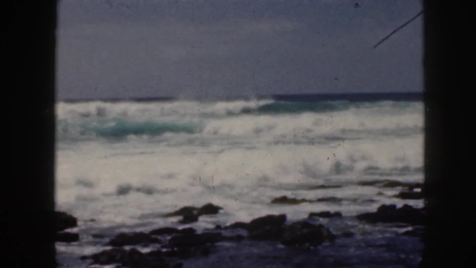 1959 Hawaii home-movie view of breaking ocean waves, rocky foreground, pale sky, and dark vertical borders.