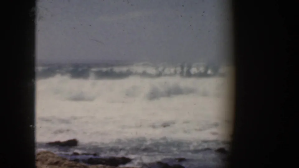 1959 Hawaii frame showing bright foaming surf across the shoreline, dark rocks below, and soft faded 8mm color.