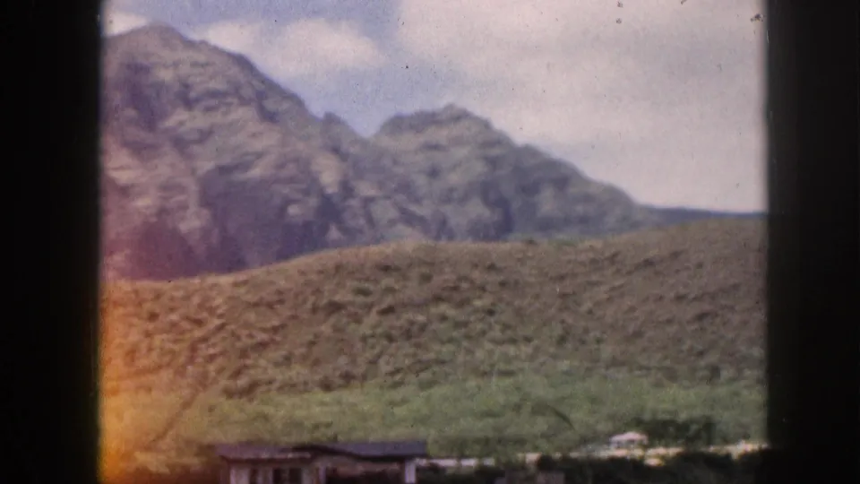 1959 Hawaii landscape view showing rocky peaks, rolling green hills, and small low structures at the lower edge of the frame.