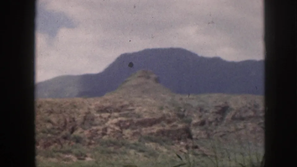 1959 Hawaii home-movie frame with a rounded foreground hill, darker mountain backdrop, and scrub vegetation under clouds.
