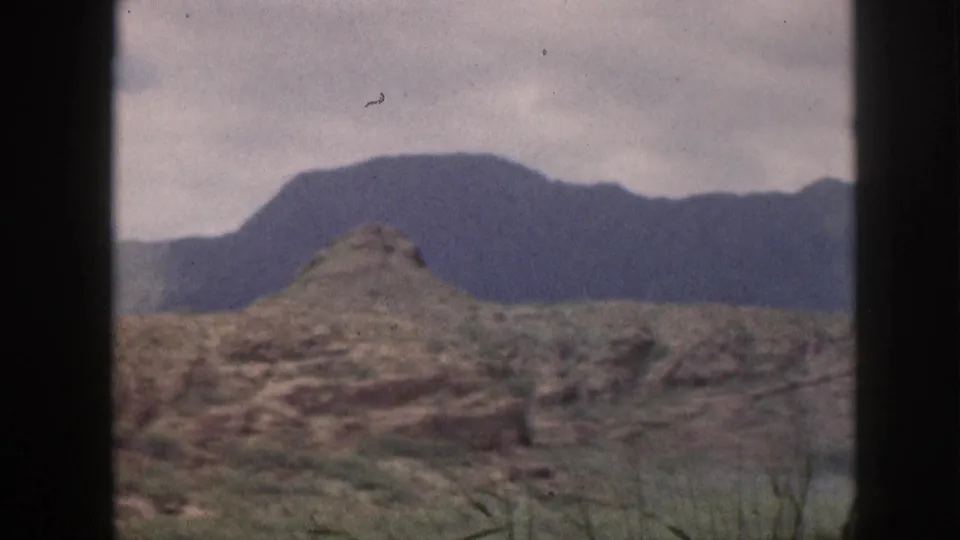 1959 Hawaii 8mm frame showing muted mountain terrain, rocky foreground, sparse vegetation, and a dark window frame.