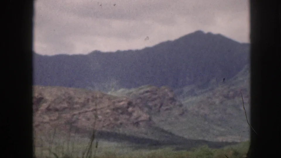 1959 Hawaii home-movie frame with distant ridgeline, rocky foreground, muted green slope, and dark window bars.
