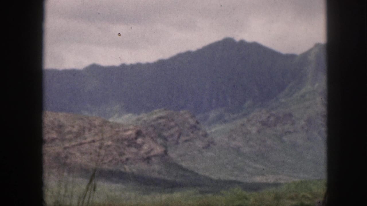 1959 Hawaii 8mm frame showing green mountain ridges, rocky lower slopes, cloudy sky, and a dark window frame in the foreground.