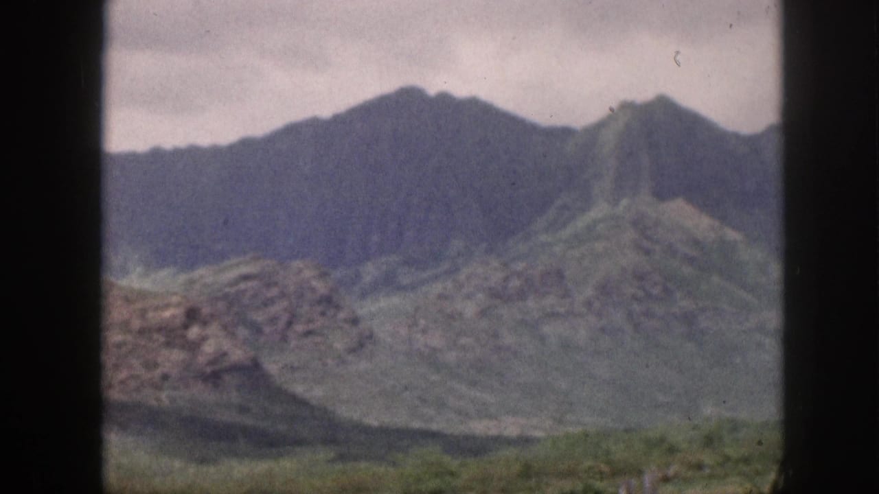 Mountain range with green slopes and rocky foreground framed by dark window bars.