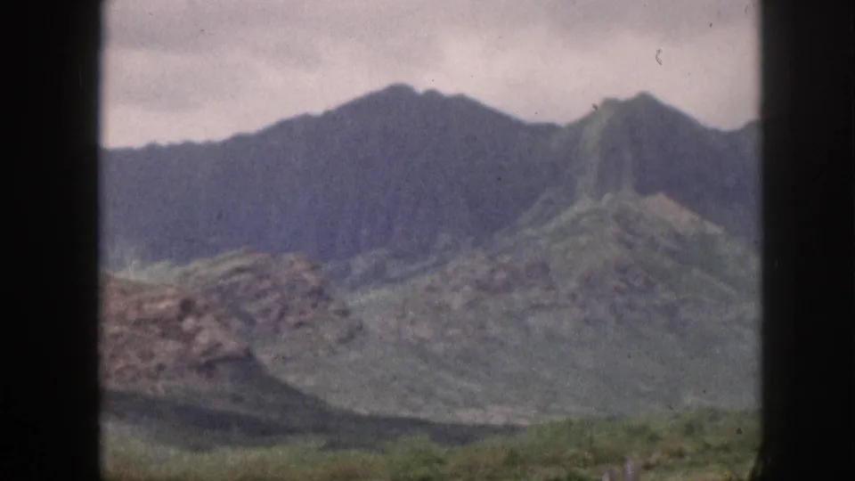 1959 Hawaii landscape frame with muted green mountains, rocky outcrops, cloudy sky, and dark window-frame edges crossing the view.