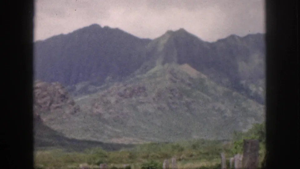 1959 Hawaii 8mm frame showing steep green mountain slopes, a rocky lower ridge, cloudy sky, and window framing at the edges.