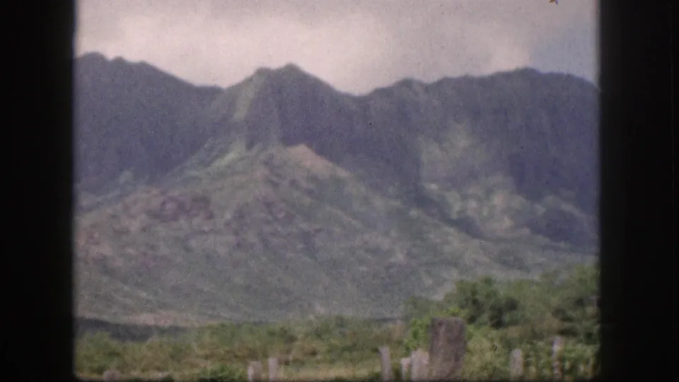 1959 Hawaii frame with green and rocky mountain slopes, cloudy sky, foreground vegetation, and dark window bars dividing the view.