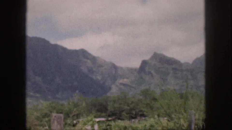 1959 Hawaii landscape view with grey mountain peaks, brush-covered foreground, cloudy sky, and a dark divider through the frame.