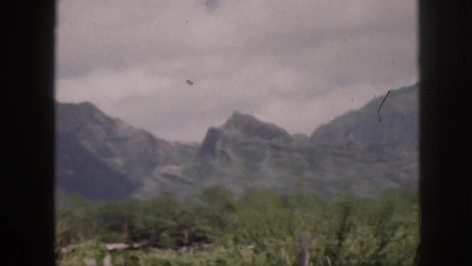 1959 Hawaii 8mm frame with distant grey mountains, low brush, cloudy sky, and dark window-like bars in the foreground.