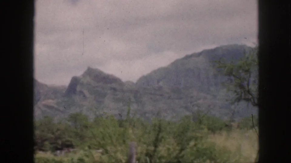 1959 Hawaii home-movie frame showing grey mountain ridges, dense valley brush, cloudy sky, and a tree at the right edge.