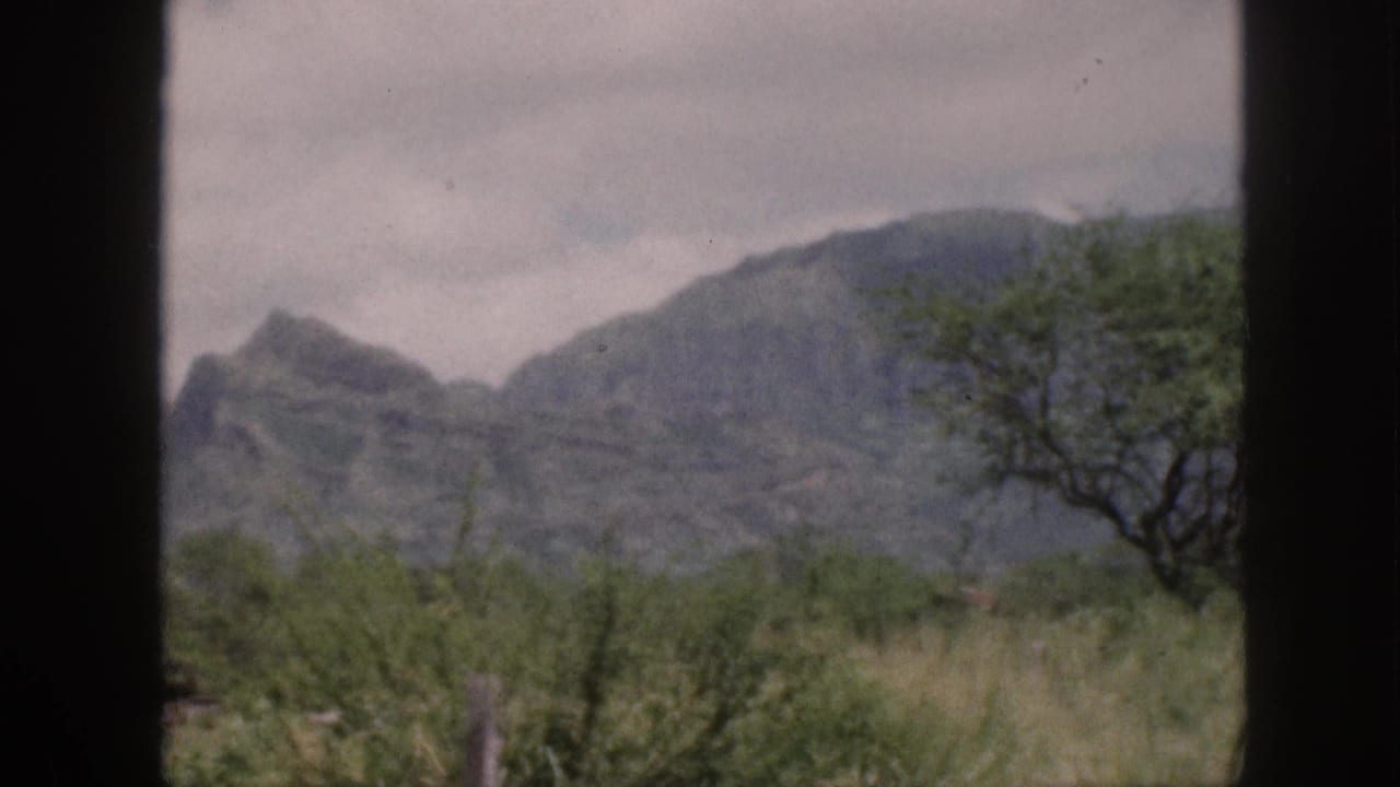 1959 Hawaii home-movie frame showing mountains, cloudy sky, roadside grass, and dark vehicle window framing.