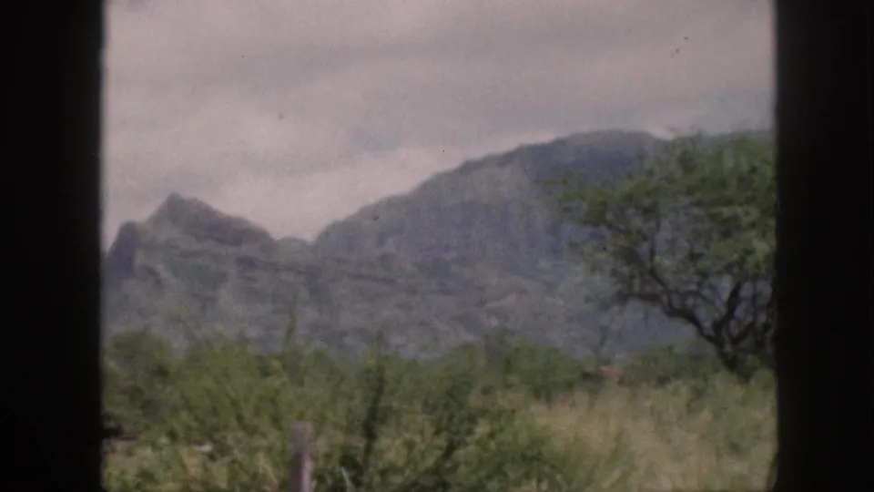 1959 Hawaii home-movie frame showing mountains, cloudy sky, roadside grass, and dark vehicle window framing.