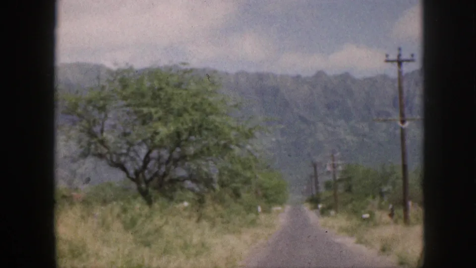 1959 Hawaii frame showing a narrow road, utility poles, roadside grass, a tree, and mountain backdrop.