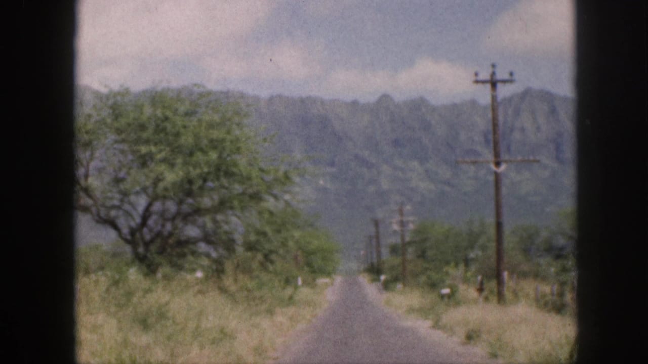 1959 Hawaii 8mm frame showing a rural road through a car window, with roadside vegetation, utility poles, and distant mountains.