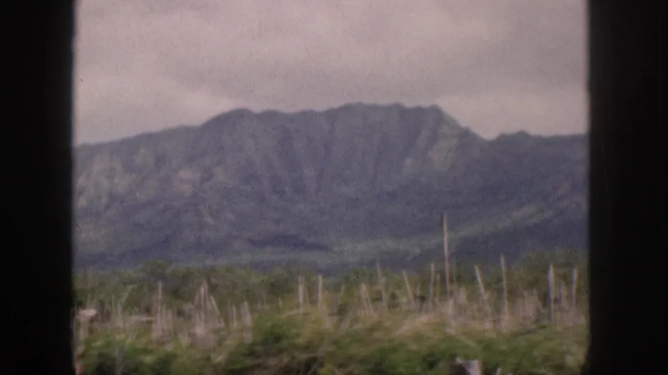 Soft 8mm view of a Hawaiian mountain ridge in 1959, framed by window bars with grasses and shrubs below.
