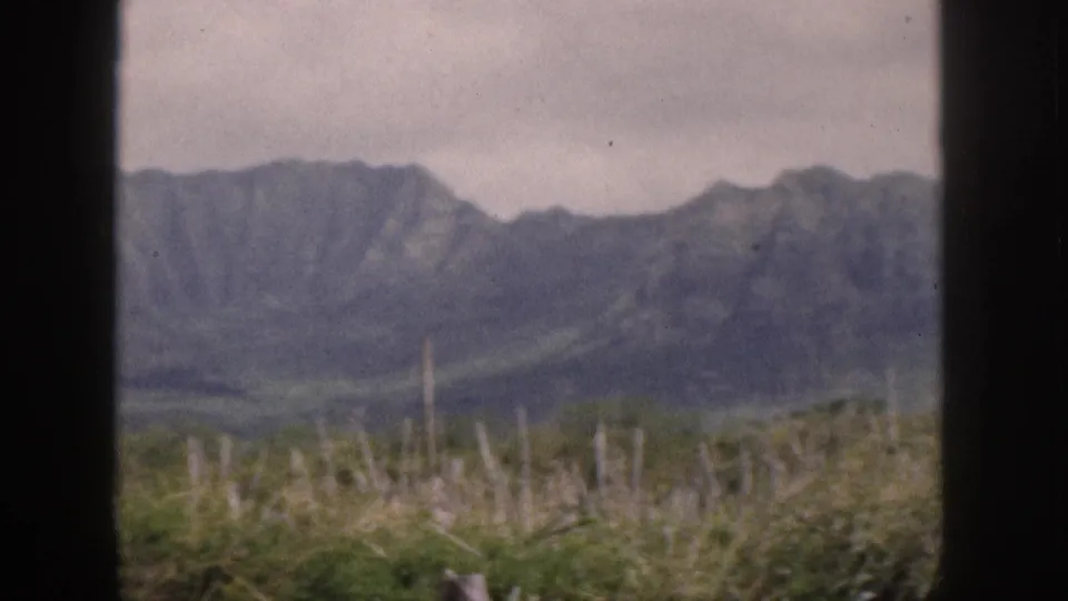 Final 1959 Hawaii frame with clouded mountain peaks, scrub vegetation, and dark window framing from the filming location.