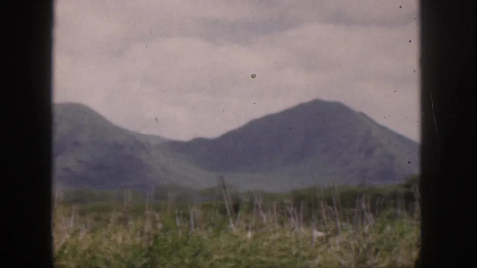 1959 Hawaii mountain landscape with soft green hills, cloudy sky, foreground grass, and visible 8mm film grain.