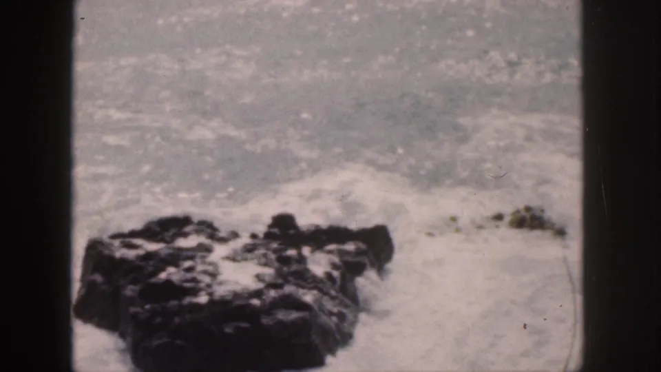 Foam and spray wash around dark coastal rocks in a 1959 Hawaii home-movie frame.