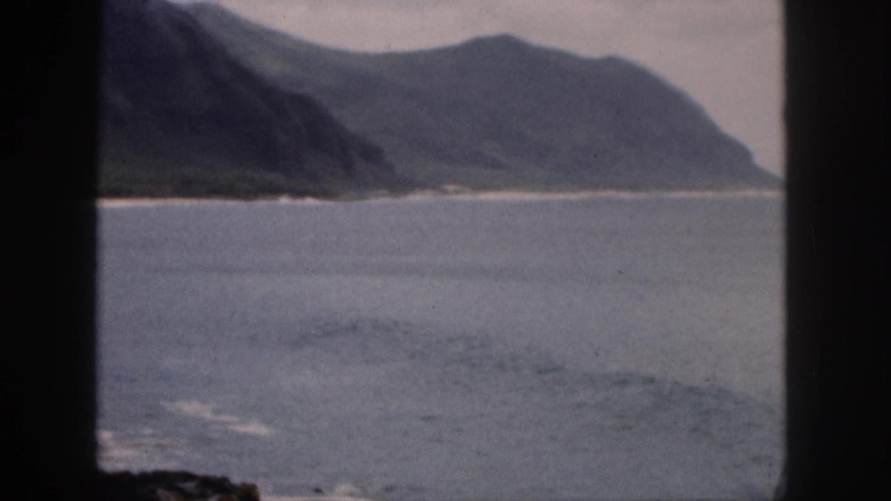 1959 Hawaii home-movie view of a coastal cove with blue water, pale shore, green mountains, and dark window-frame bars.