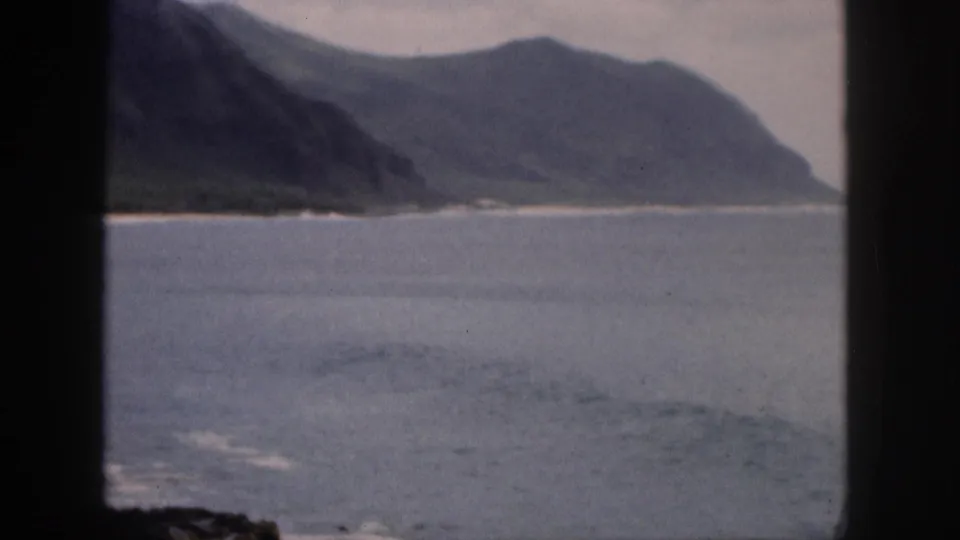 1959 Hawaii home-movie view of a coastal cove with blue water, pale shore, green mountains, and dark window-frame bars.
