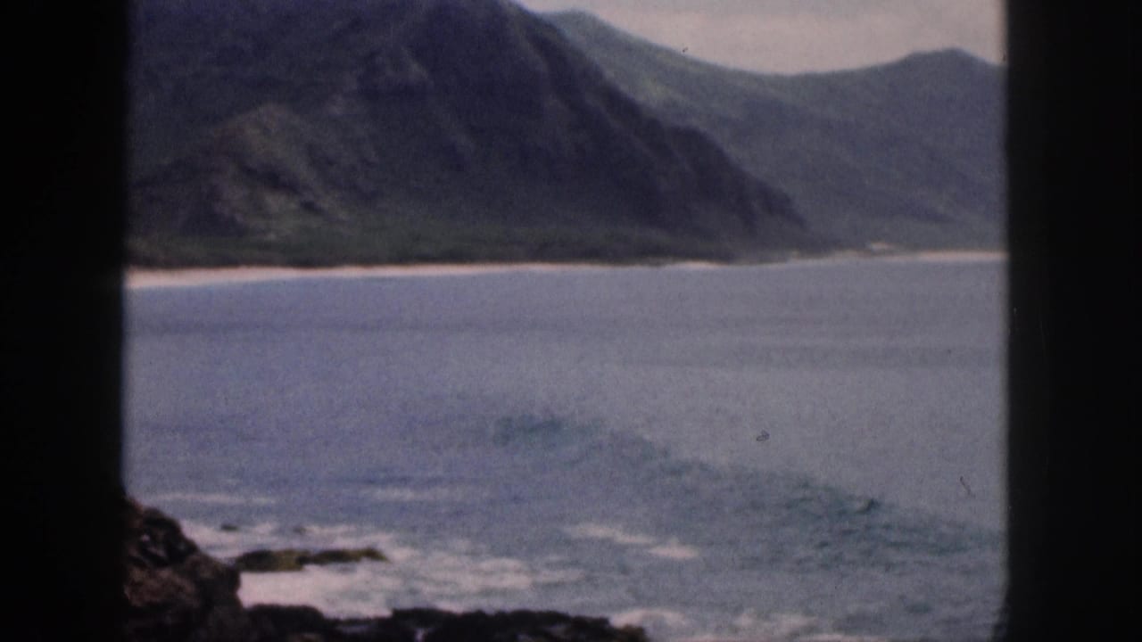 Mountain coastline and small surf viewed across blue water through a dark frame.