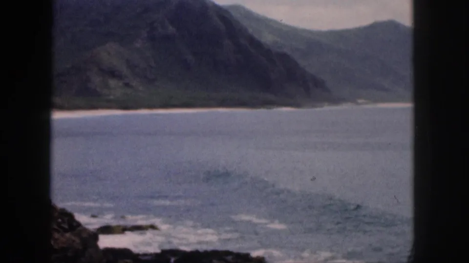 1959 Hawaii 8mm frame showing a mountain-backed shoreline, low surf, blue water, and a dark window divider in the foreground.