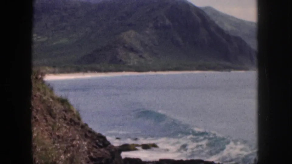 1959 Hawaii coastal view with rocks in the foreground, small waves in the cove, sandy shore, and steep green mountains beyond.