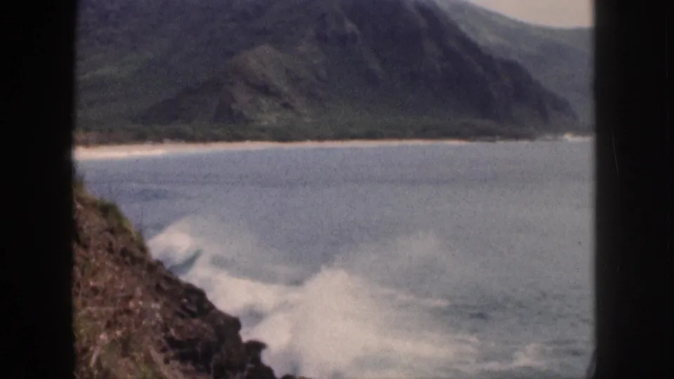 1959 Hawaii home-movie frame of white surf breaking beside dark rocks, with blue cove water and a mountain shoreline in the distance.