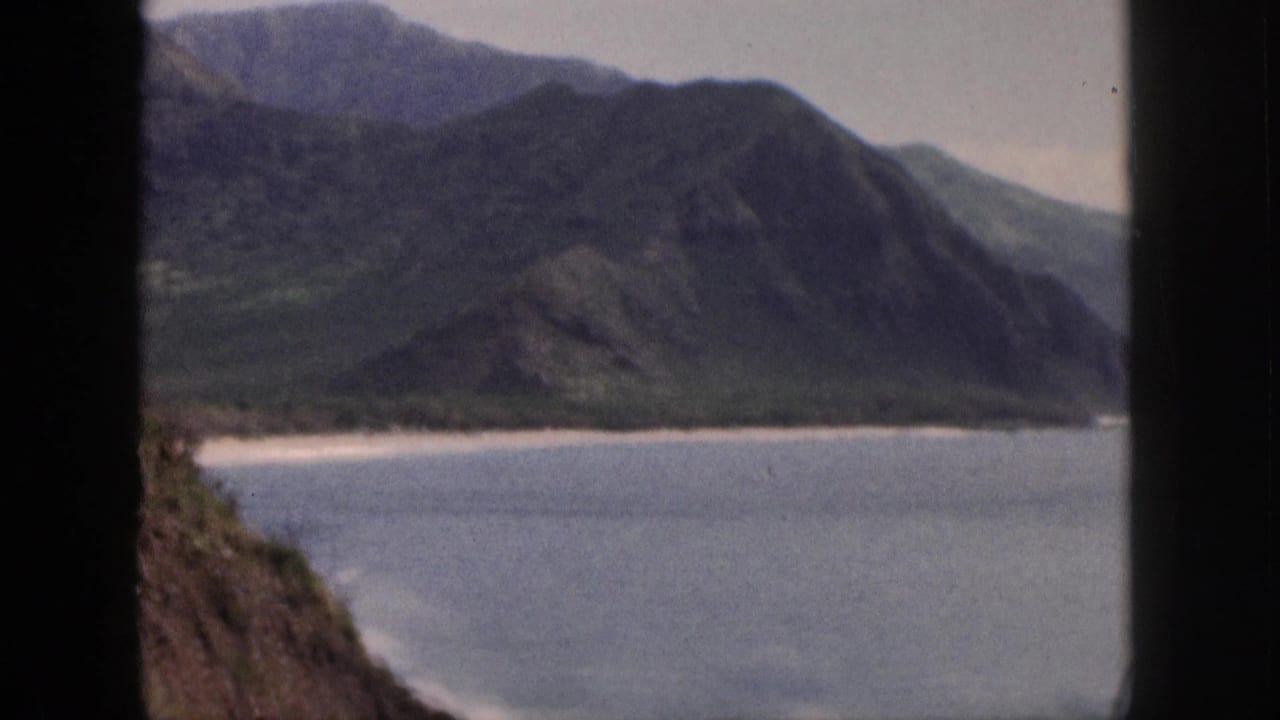 1959 Hawaii home-movie frame showing a calm bay and steep green mountains from a roadside viewpoint.