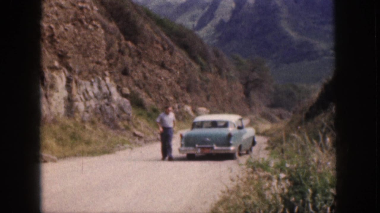 Adult man stands near a turquoise-and-white 1950s sedan on a narrow Hawaii cliff road, with mountains behind.