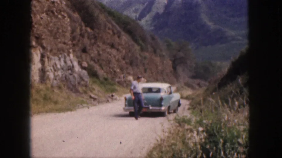 A man stands by a turquoise-and-white sedan on a winding Hawaii mountain road in this 1959 home-movie frame.