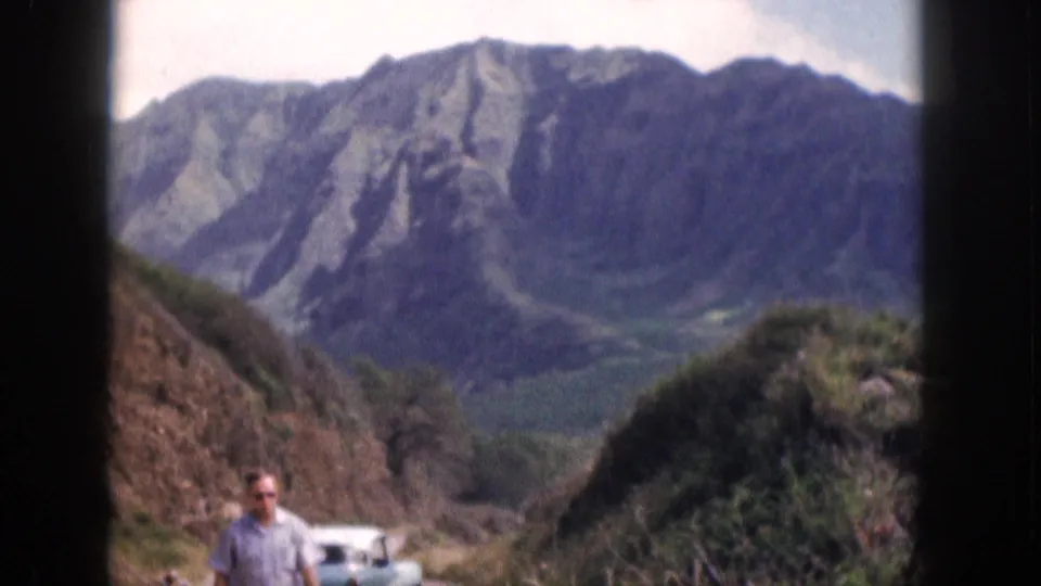 1959 Hawaii frame with an adult man near a parked sedan at a mountain roadside overlook.