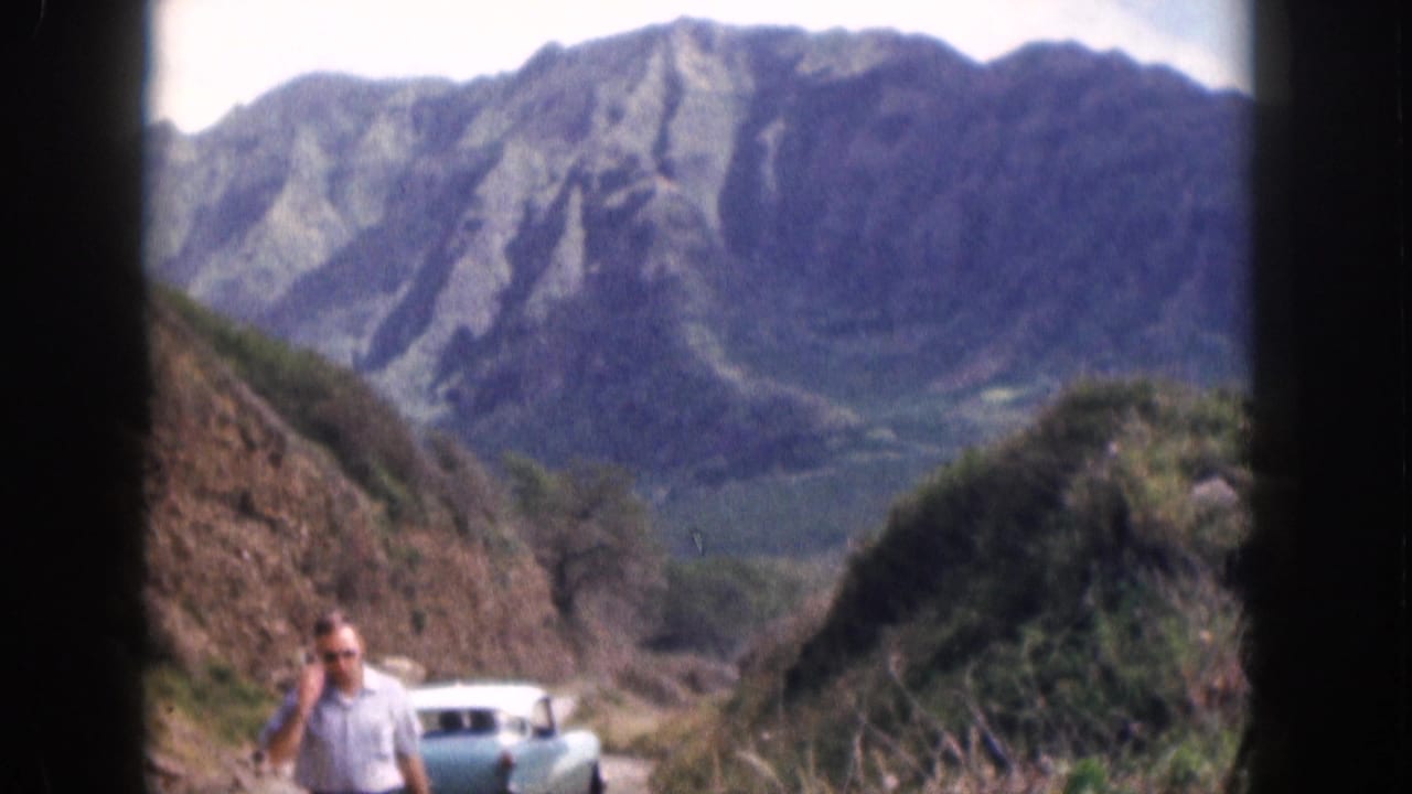 1959 Hawaii home-movie frame showing a man in sunglasses on a narrow mountain road with a turquoise sedan and ridged mountains behind him.