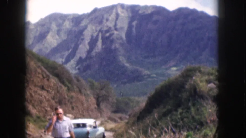 1959 Hawaii home-movie frame showing a man in sunglasses on a narrow mountain road with a turquoise sedan and ridged mountains behind him.