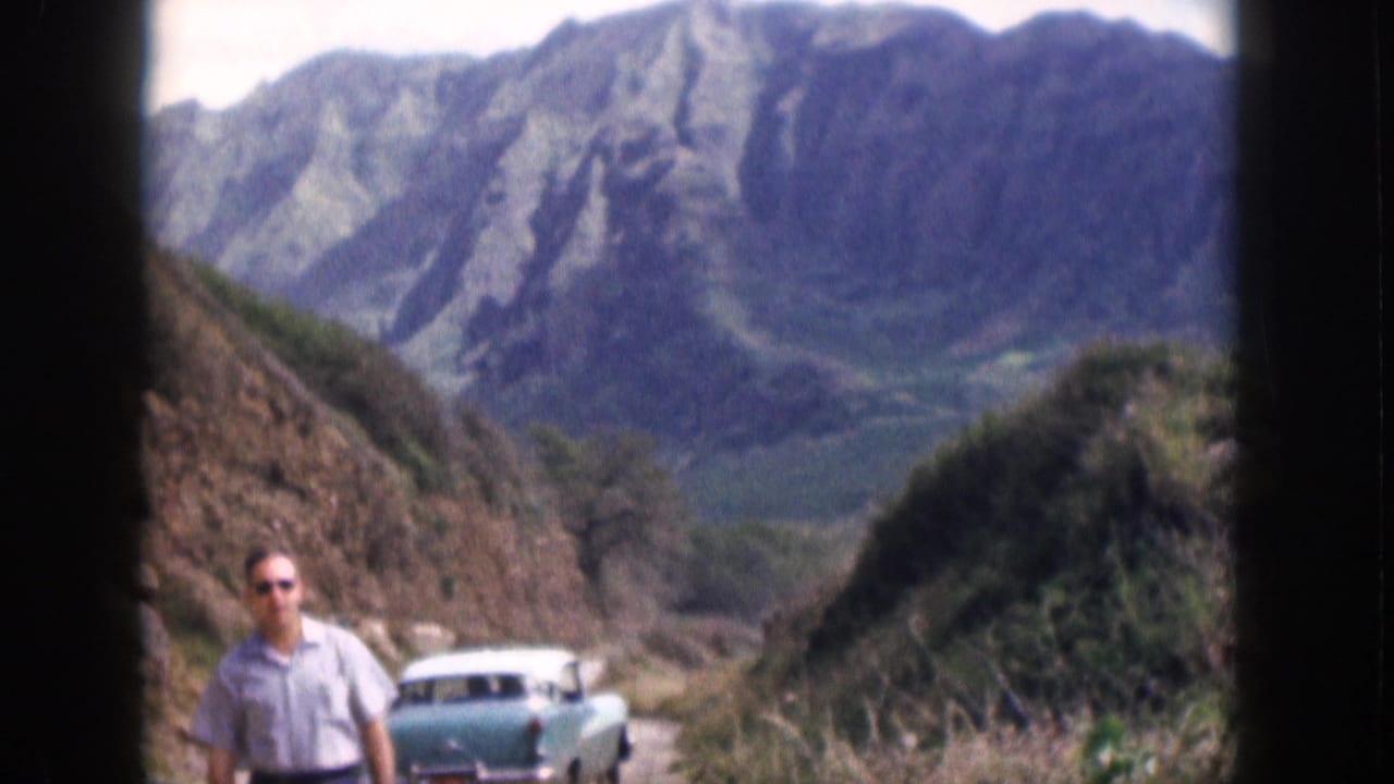 Man stands near a winding road with a turquoise sedan and mountains in the background.