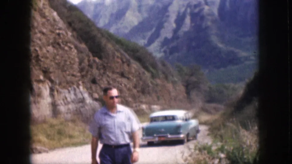 1959 Hawaii 8mm frame showing a man walking toward the camera on a curving mountain road with a turquoise sedan behind him.