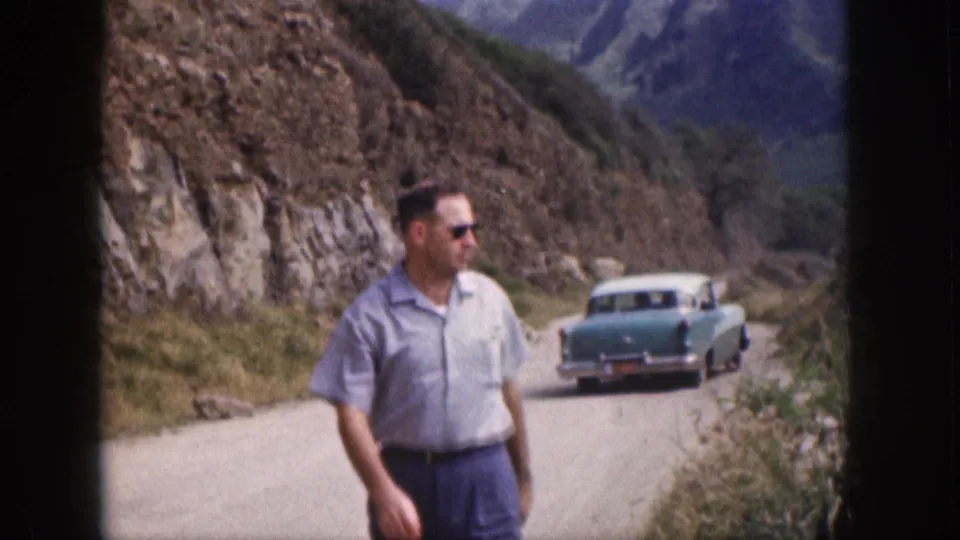 1959 Hawaii home-movie frame showing the man closer to camera, with a parked turquoise classic sedan and rugged mountains beyond the road.