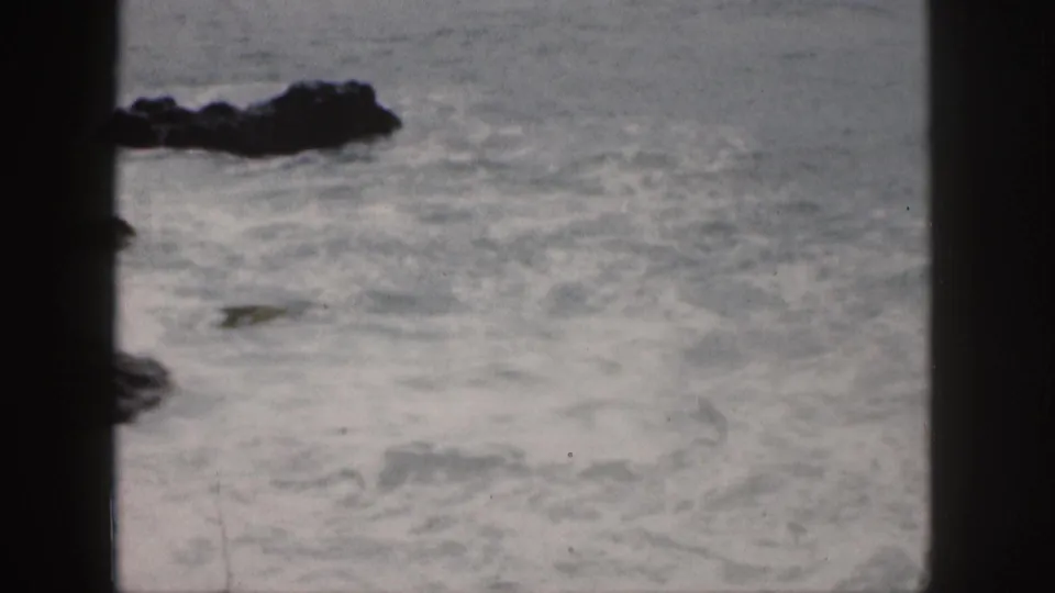 1959 Hawaii 8mm frame showing foamy surf around a dark shoreline rock, with grain and faded color visible.