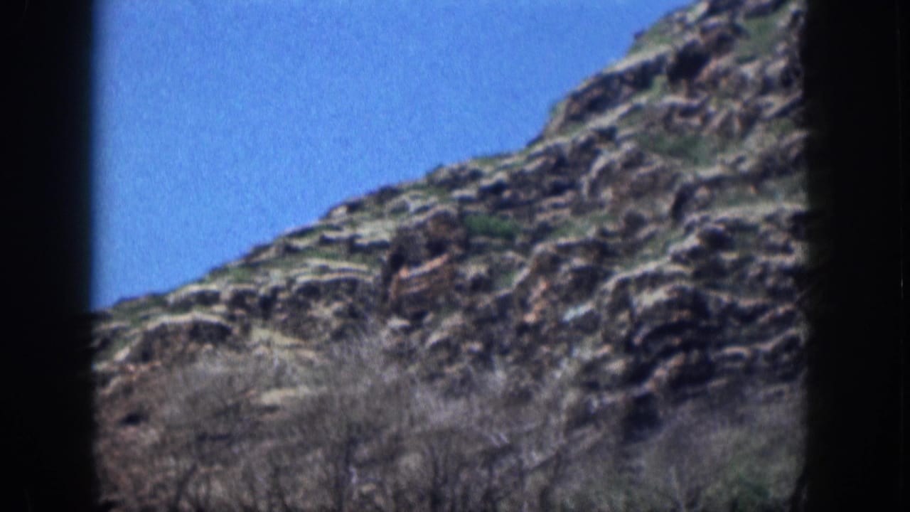 Rocky mountainside with dry brush below a clear blue sky, bordered by dark film edges.