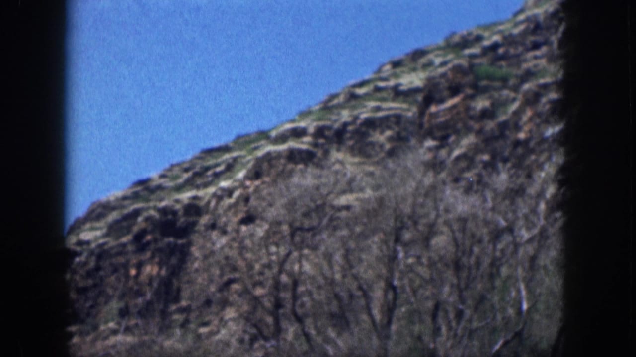 Rugged hillside with sparse shrubs and blue sky, partially framed by dark vertical borders.