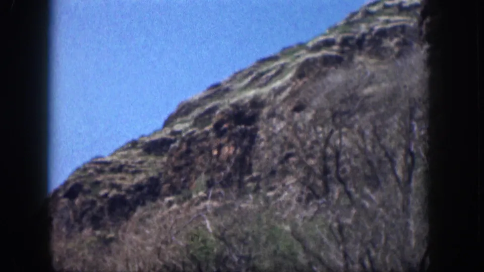 1959 Hawaii home-movie still showing a diagonal rocky mountainside, dry grasses, sparse shrubs, and saturated blue sky.