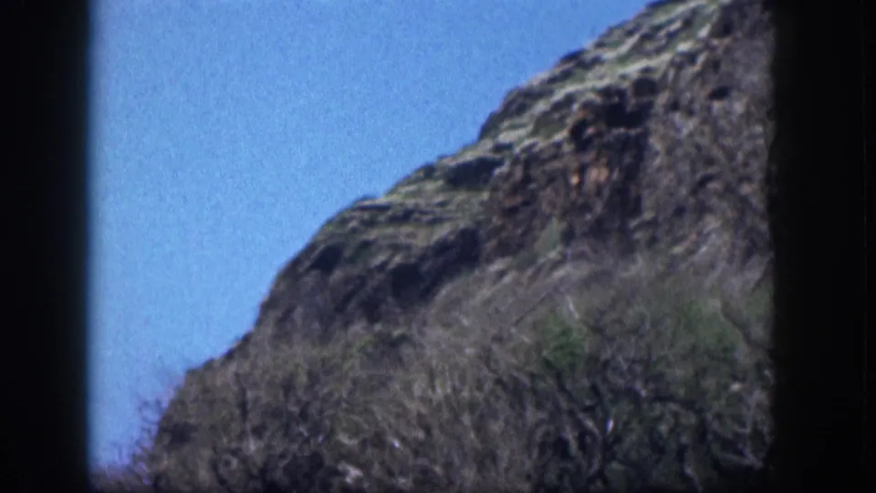 1959 Hawaii frame showing a rocky mountainside edge, dry scrub, clear blue sky, and dark vertical film-frame borders.