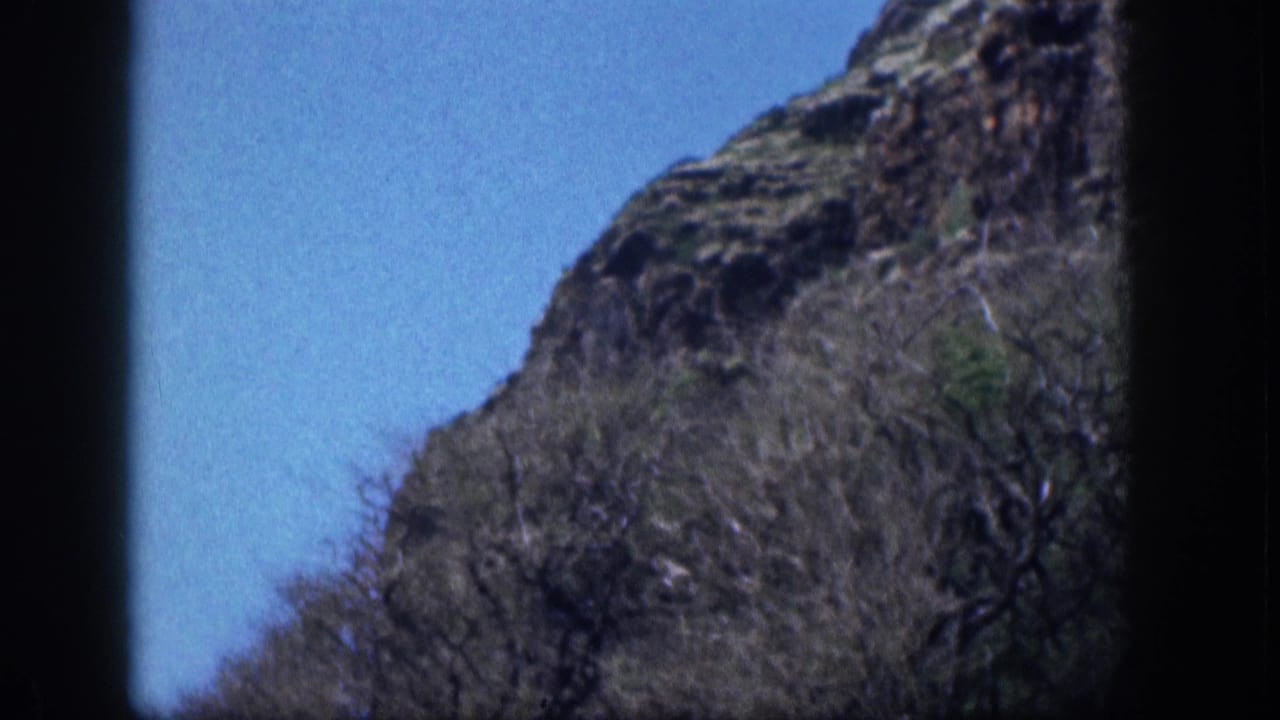 Steep brush-covered mountainside beneath blue sky with dark 8mm frame borders.