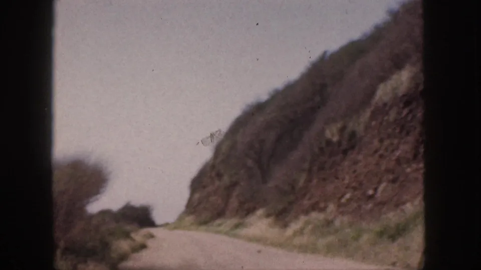 1959 Hawaii 8mm frame showing a pale winding road at the base of rocky slopes, with sparse roadside vegetation and sky.