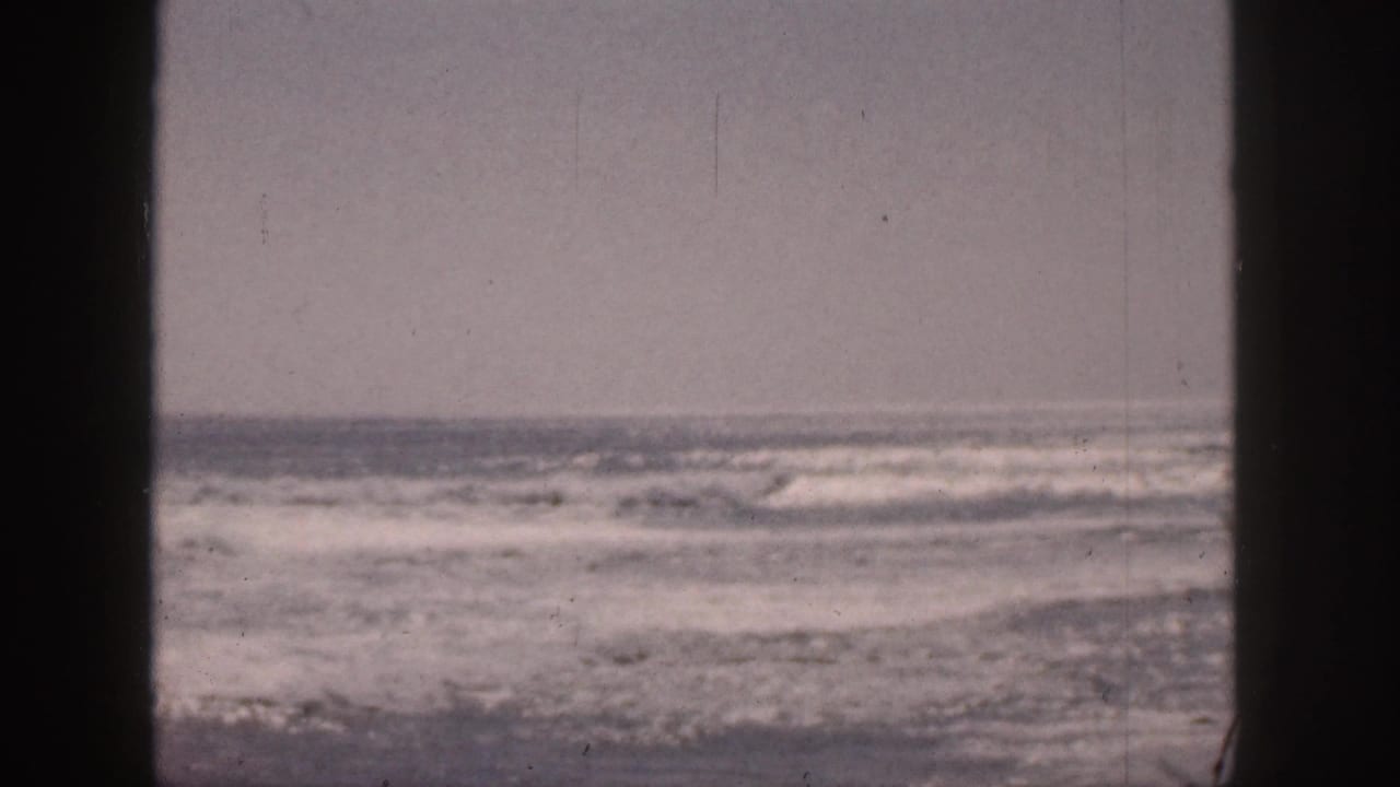 1959 Hawaii 8mm frame showing rough surf and whitecaps viewed through dark window-like vertical bars beneath an overcast sky.
