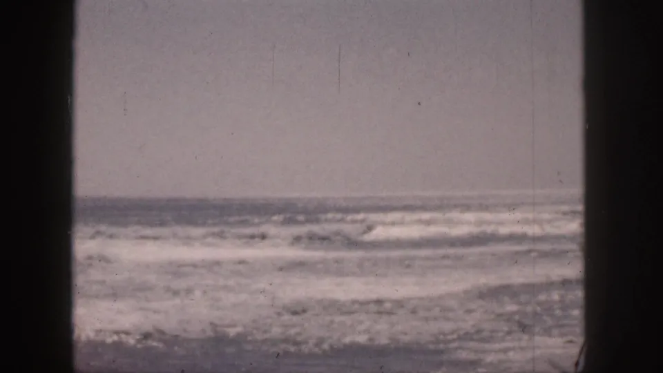 1959 Hawaii 8mm frame showing rough surf and whitecaps viewed through dark window-like vertical bars beneath an overcast sky.