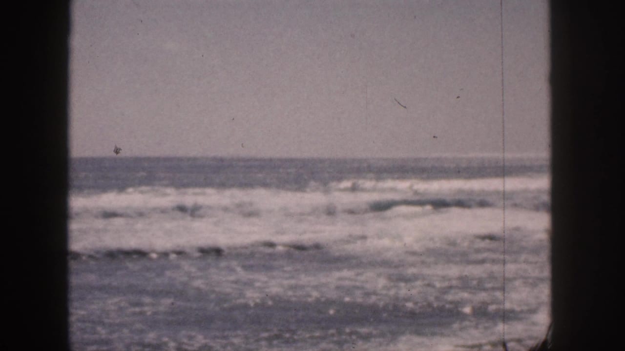 1959 Hawaii home-movie frame with rolling surf, a low horizon, muted color, and a dark vertical divider through the view.