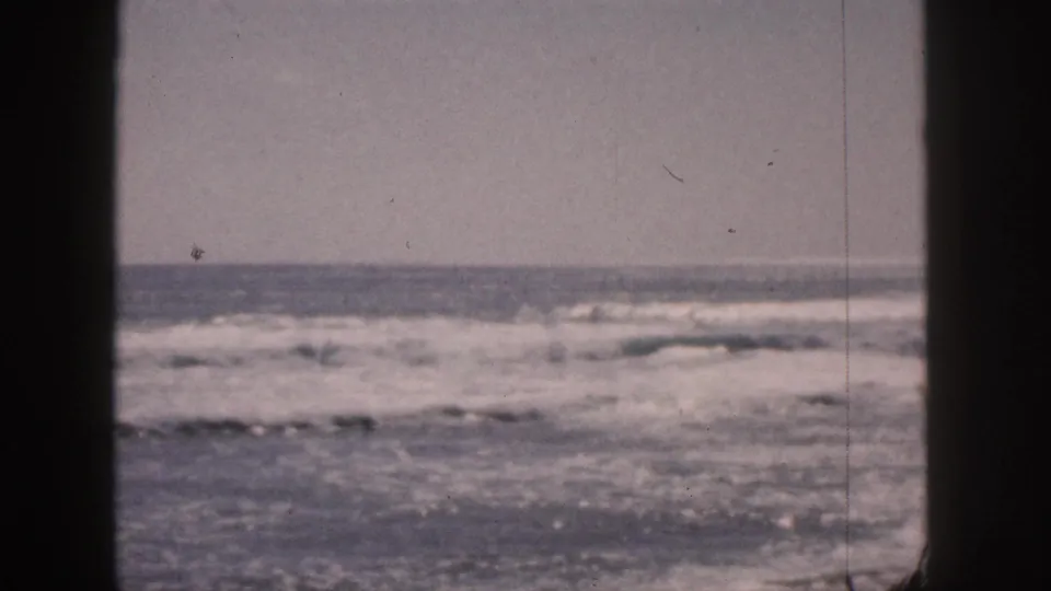 1959 Hawaii home-movie frame with rolling surf, a low horizon, muted color, and a dark vertical divider through the view.