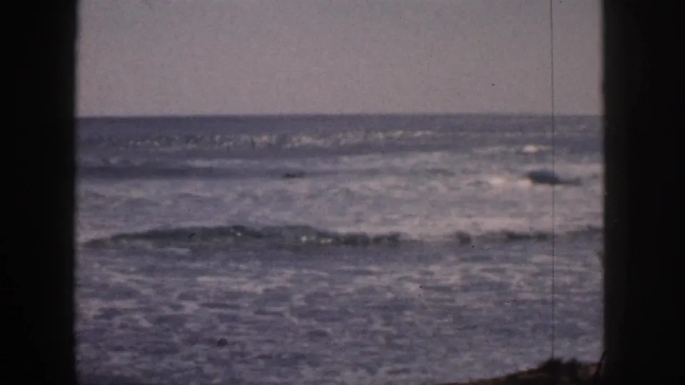 1959 Hawaii 8mm still showing calmer ocean water, low surf, pale sky, and dark vertical framing along the left and center.